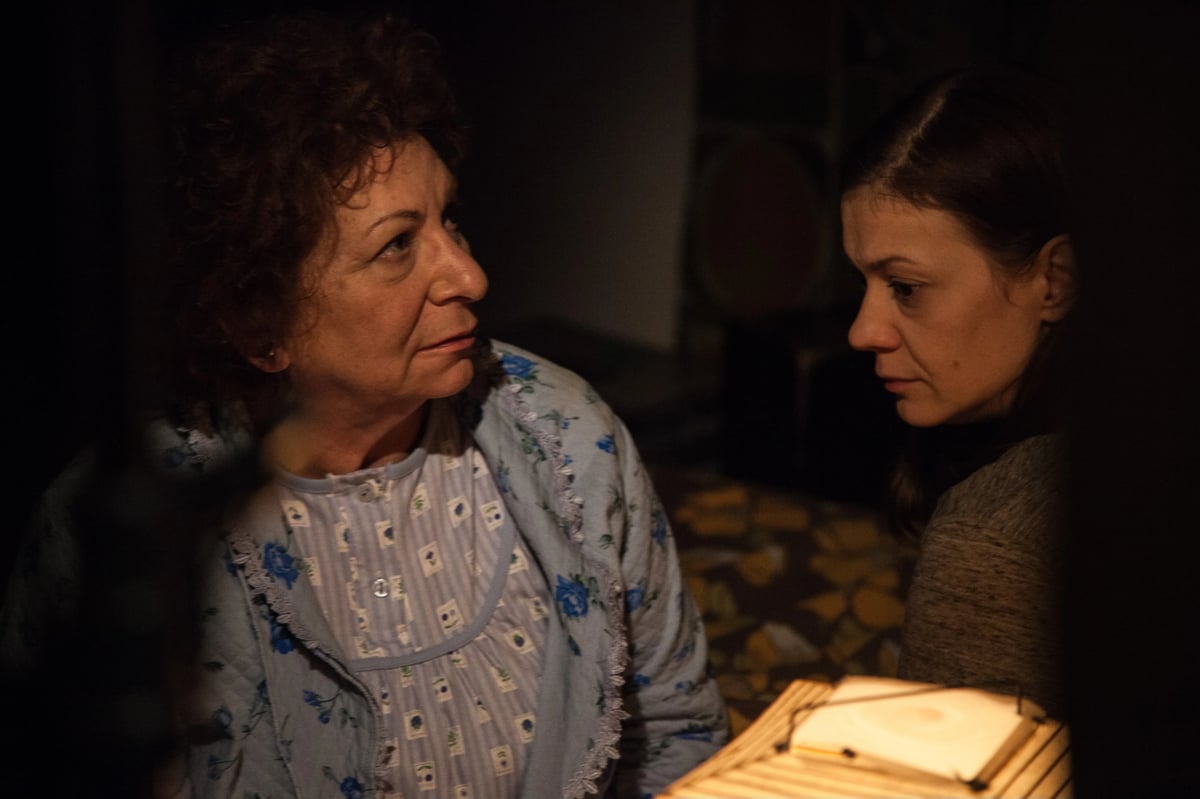 Two women in a dim, warm-lit interior scene looking at each other while seated close together.