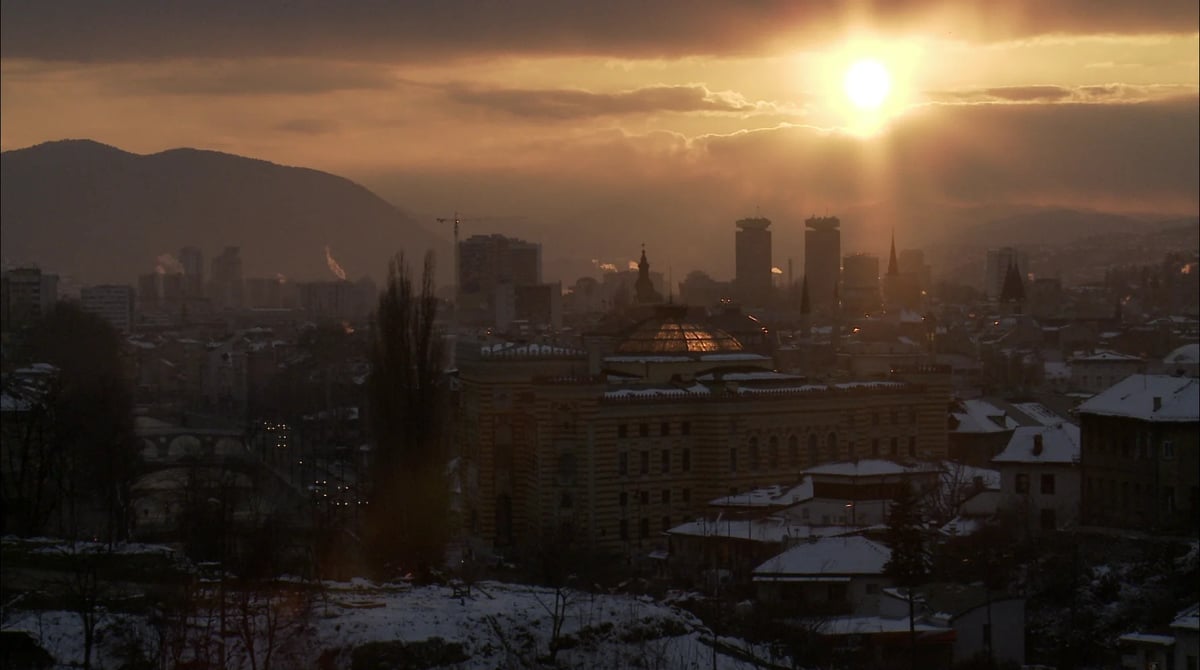Wide skyline view at sunset with low sun behind clouds, hills in the distance, and winter rooftops below.