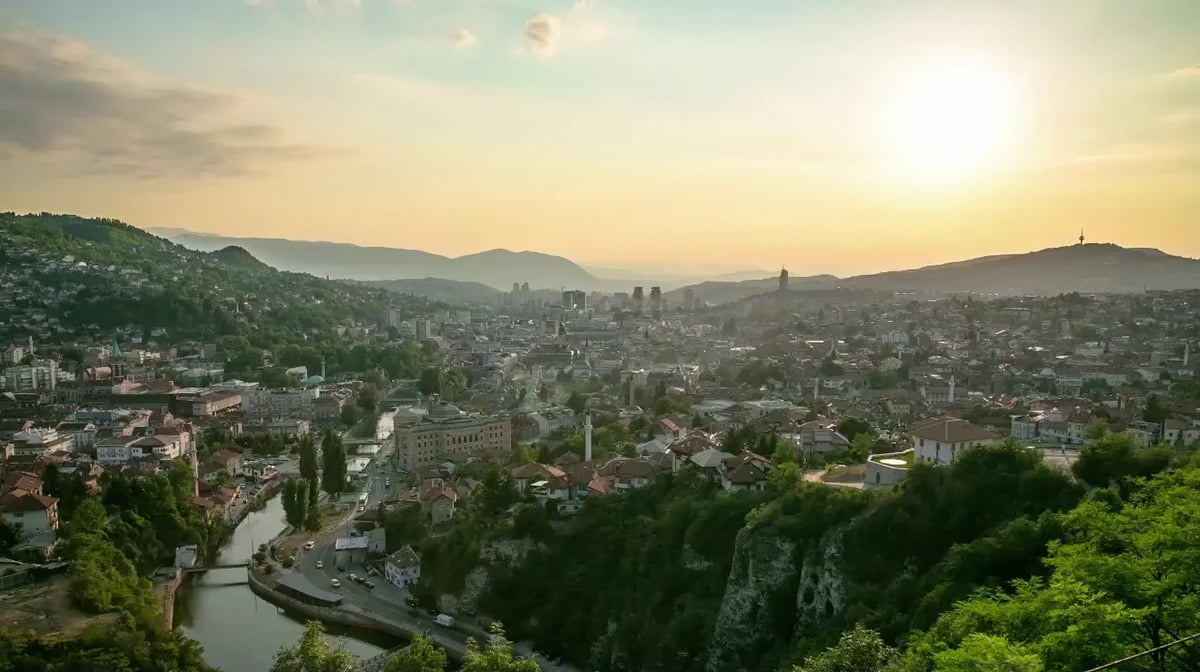 Wide panoramic sunset cityscape with a river through a dense hillside town and mountains in the background.
