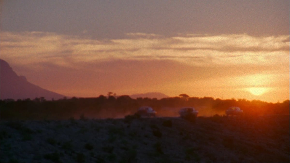Sunset over a desert landscape with distant mountains and silhouettes of vegetation in the foreground.