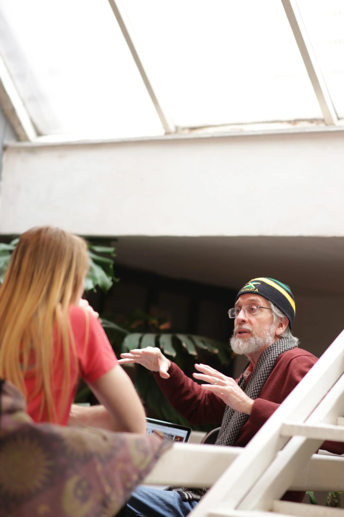 An older bearded man wearing a beanie speaks during an interview while a blonde woman in red gestures nearby; laptop and studio setup visible.