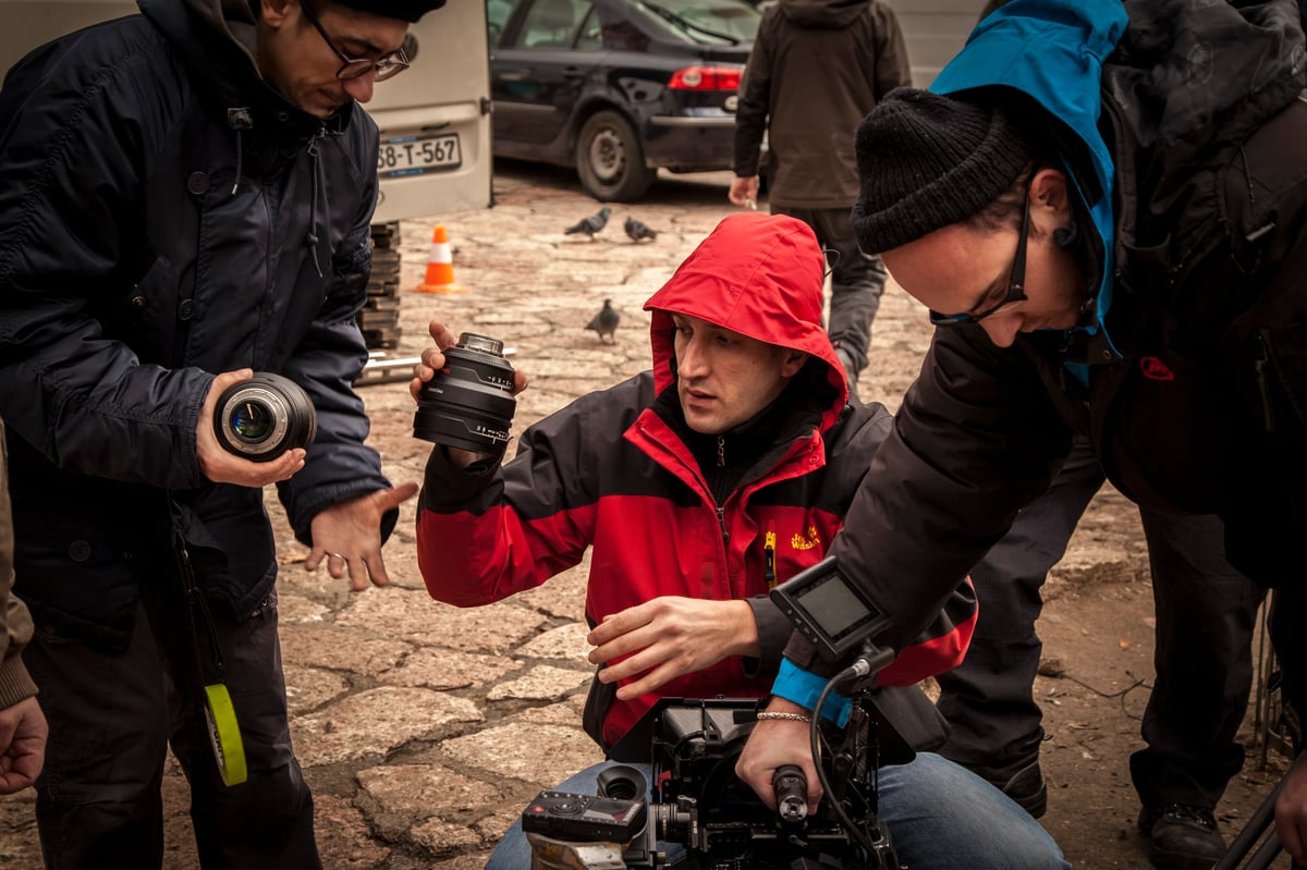 Film crew members on a snowy outdoor set using cameras and a monitor, with vehicles and crew in the background.
