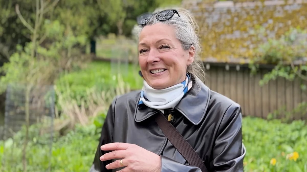 Smiling woman in a black leather jacket and white scarf standing outdoors in a garden, with sunglasses resting on her head