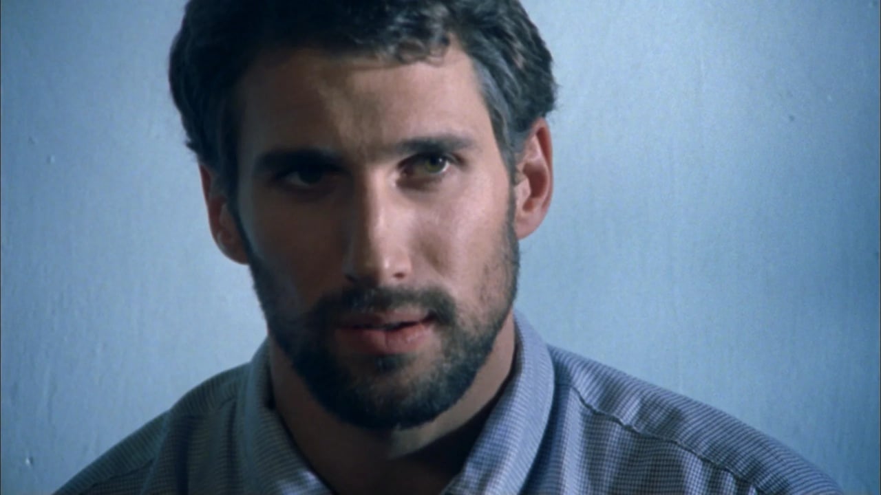 Close-up portrait of a man with short dark hair and a trimmed beard looking toward the camera against a plain blue background.