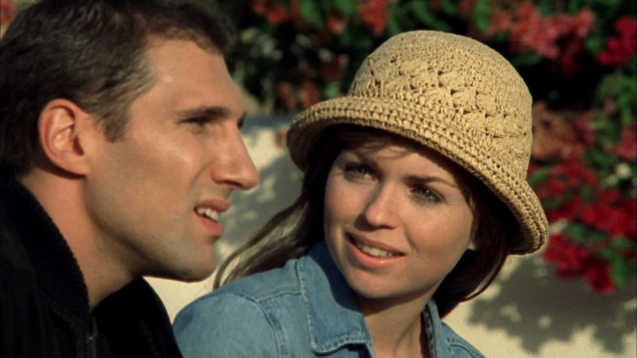 Two smiling people outdoors; one wearing a straw hat, the other in profile, in a soft-focus garden background.