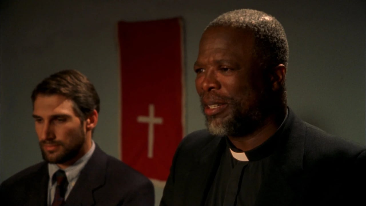 Two men in suits standing indoors, with a red flag featuring a white cross in the background.