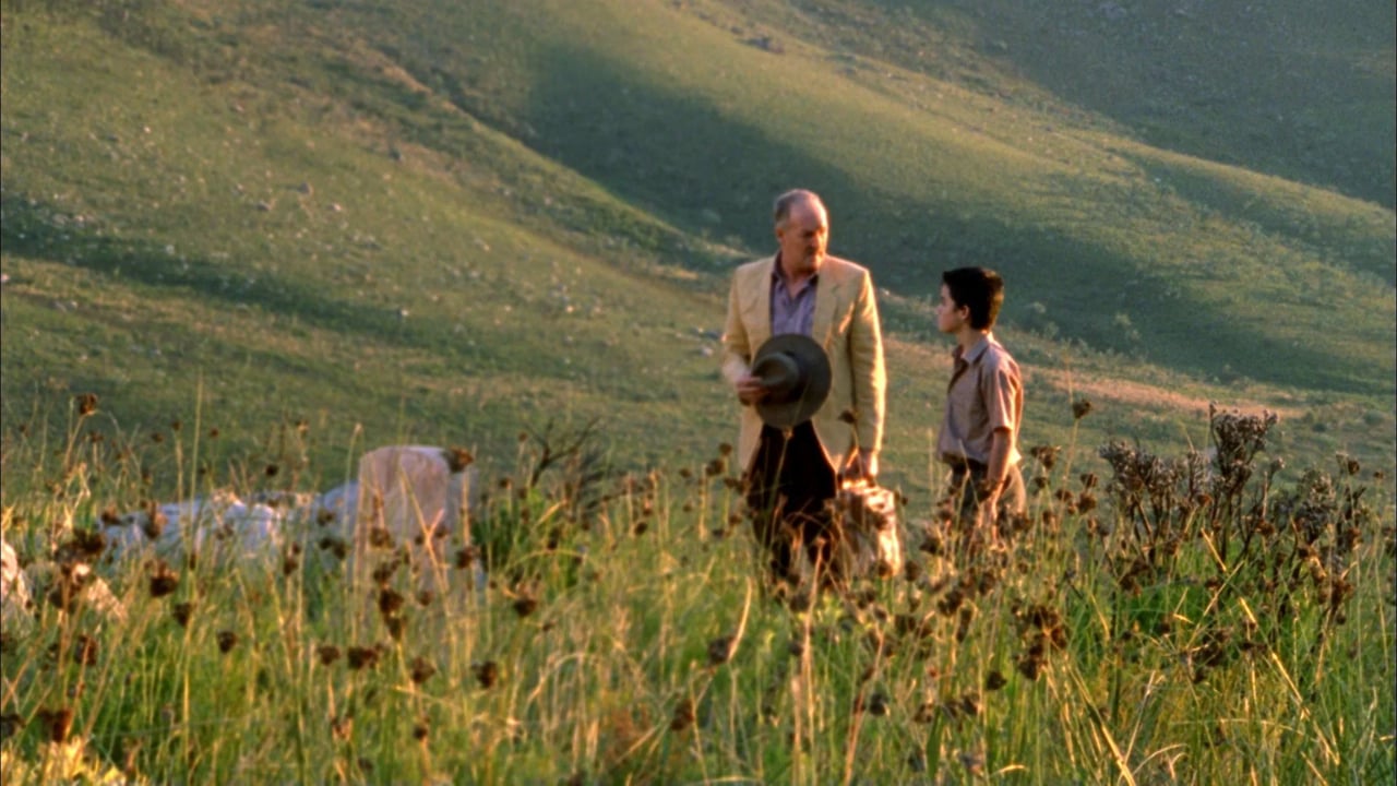 A man and a woman talk while standing in tall grass with hills in the background.