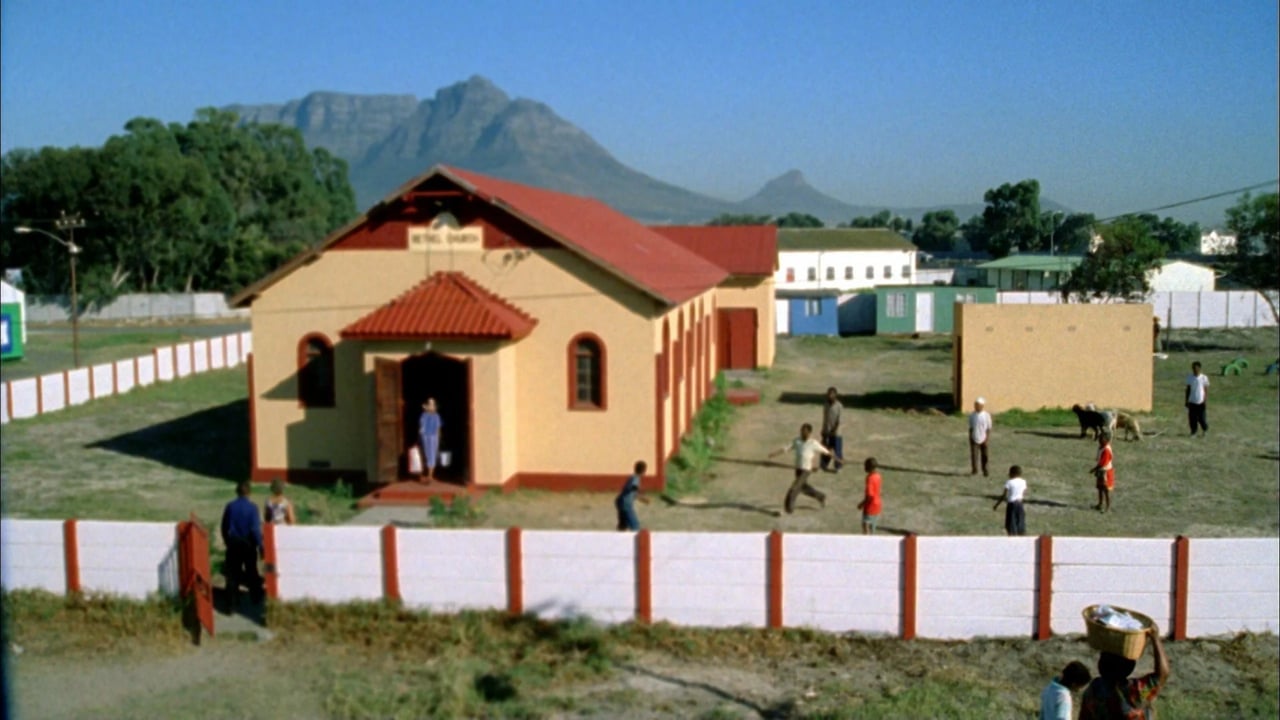 Wide outdoor scene with a red-roof building and people playing in a grassy yard, with mountains in the distance.