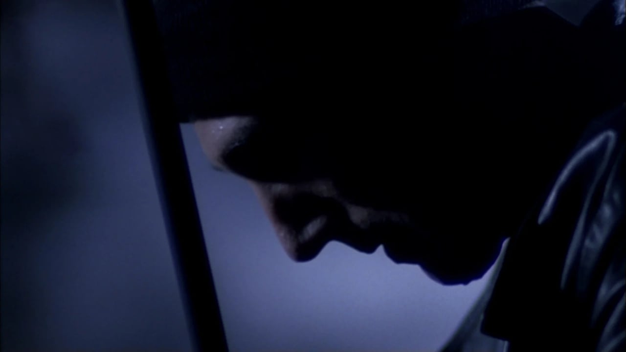 Close-up of a shadowed face in a dim blue-lit environment, partially obscured by darkness.