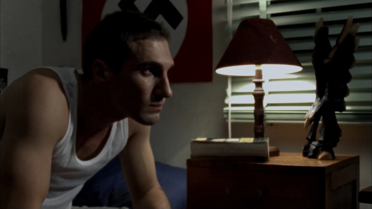 Side-profile portrait of a woman in a room with a lit lamp and a handgun on a table in the background.