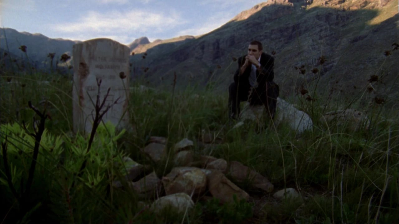 A lone man stands among tall grass in a mountain valley at dusk, with hills and clouds in the distance.