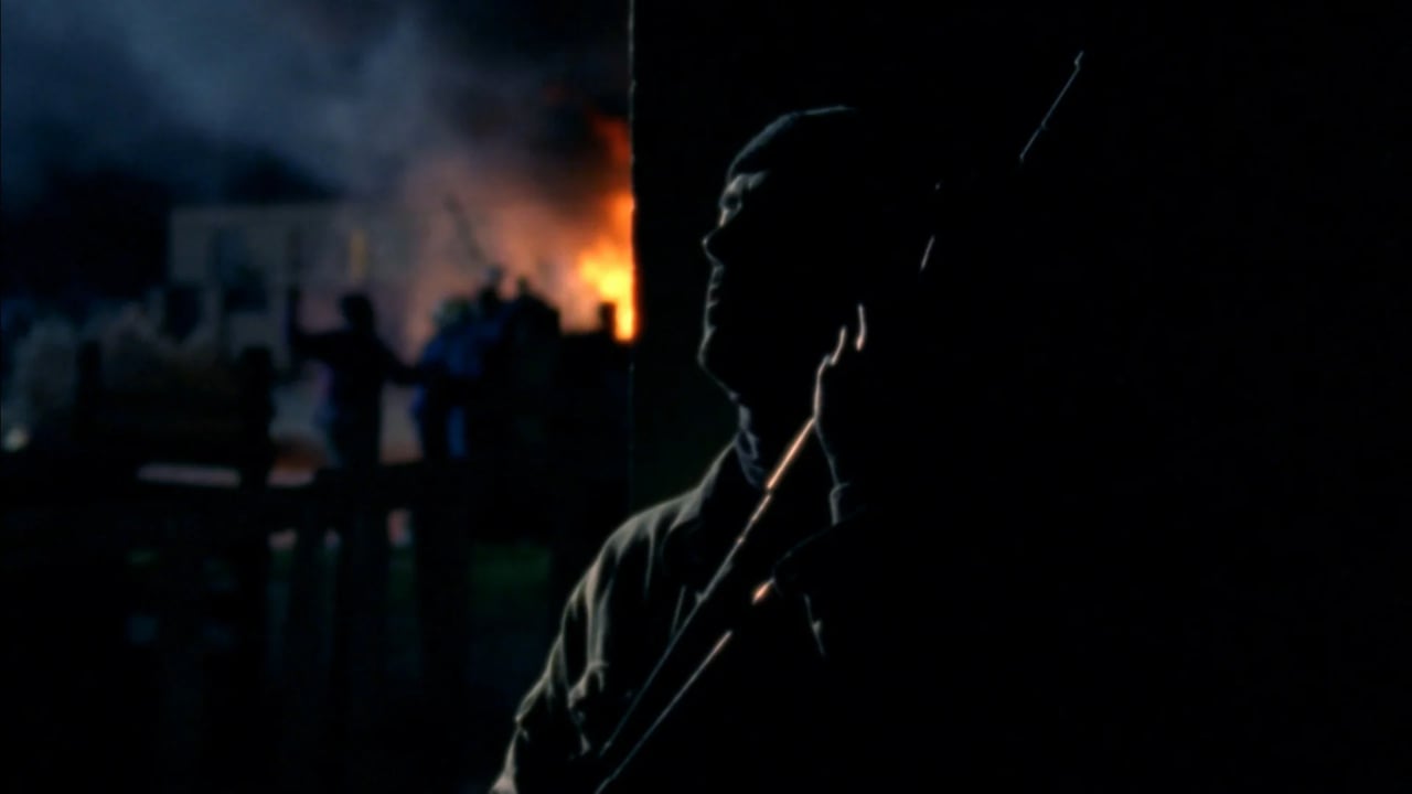 Dark cinematic image of a person’s face and profile silhouette in the foreground, with an orange fire and building in the background at night.