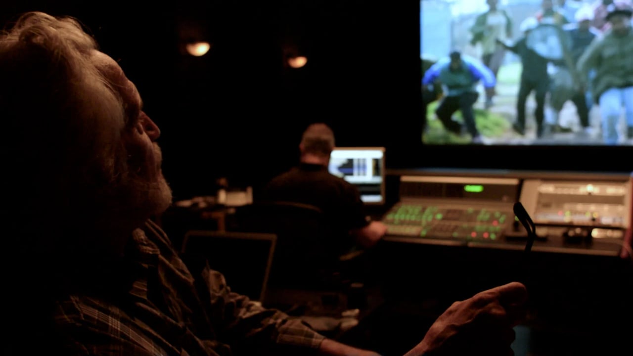 In a dark editing room, a person in the foreground watches a monitor while another person appears blurred in the background.