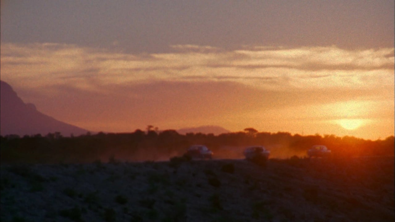 Sunset over a desert landscape with distant mountains and silhouettes of vegetation in the foreground.