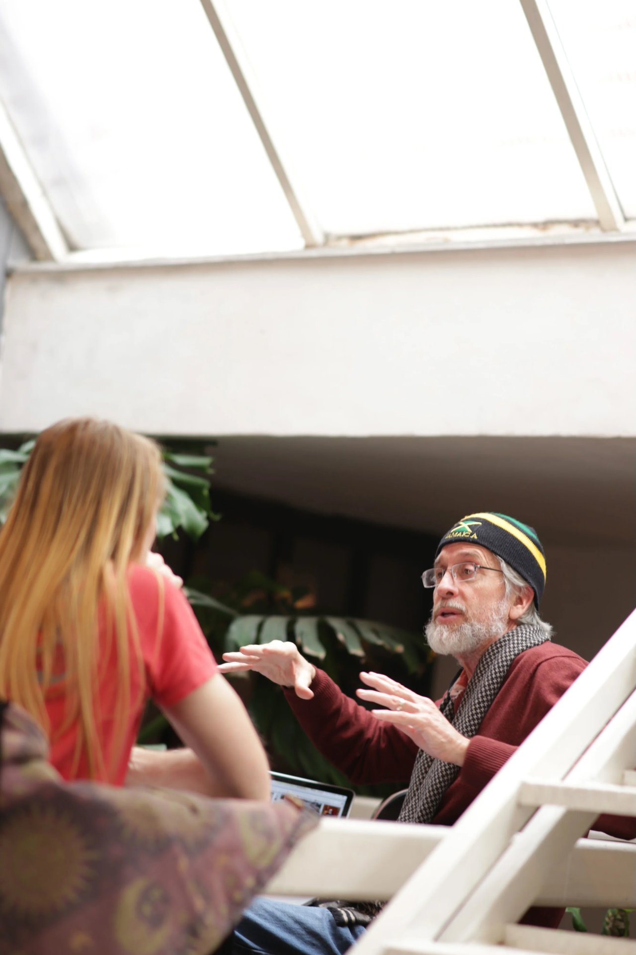 An older bearded man wearing a beanie speaks during an interview while a blonde woman in red gestures nearby; laptop and studio setup visible.