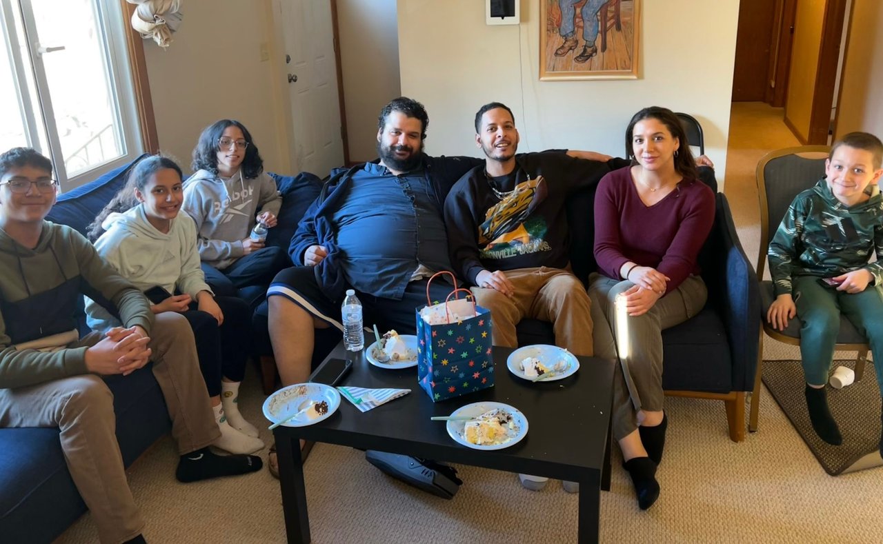 A group of seven people sitting in a living room for a casual celebration, with plates and snacks on a coffee table.
